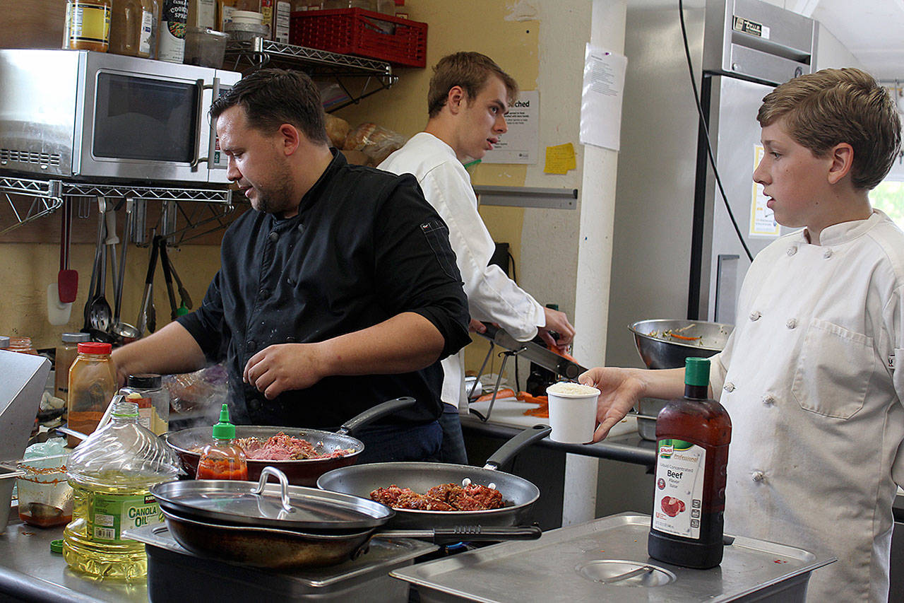 David Phillips, left, leads a crew of young cooks at The South Whidbey Commons Coffeehouse Bookstore in Langley. He created a themed menu this summer for the Island Shakespeare Festival. Caelan Boyd, right, waits to hand Phillips rice while Zakris Pierson slices carrots.(Photo by Patricia Guthrie/Whidbey News Group)